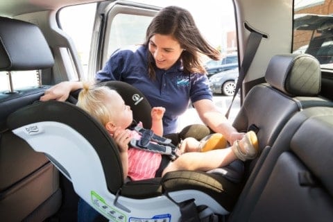 A woman securing a young child in a car seat in the backseat of a large vehicle.