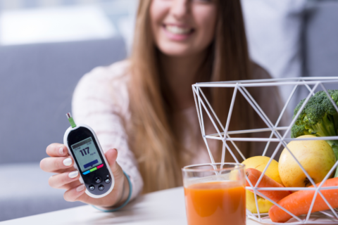 Woman holding glucose monitor at a table with fruits and veggies on it.