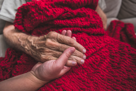 Elderly person covered with a red blanket holding the hand of a younger person.