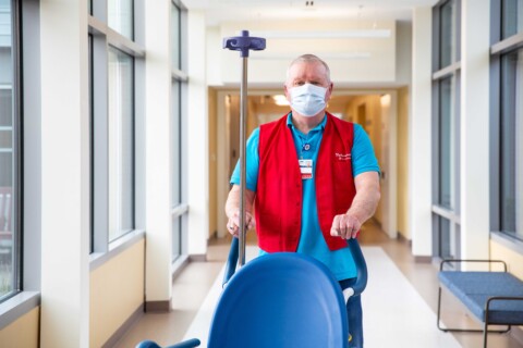 Male volunteer pushes an empty wheelchair through hospital hallway.