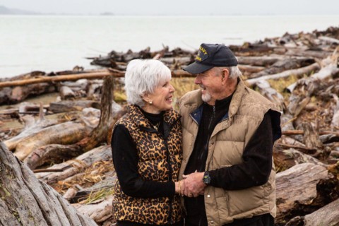 Older couple Fred and Cheri Stilwell standing on a beach covered in driftwood in the rain, smiling at each other and holding hands.