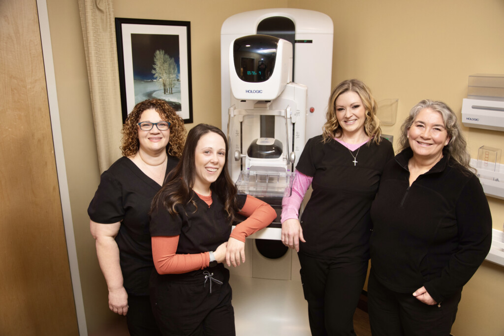 Five mammogram techs in scrubs stand in front of mammography equipment.