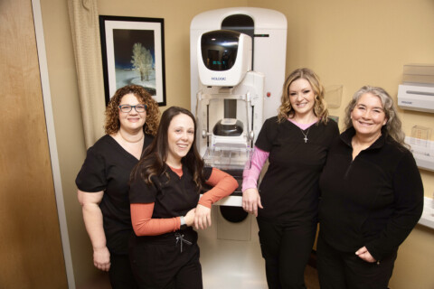 Five mammogram techs in scrubs stand in front of mammography equipment.