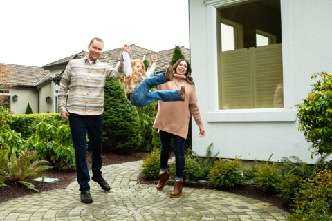 A parents  outside their PNW home swinging their daughter by the arms.