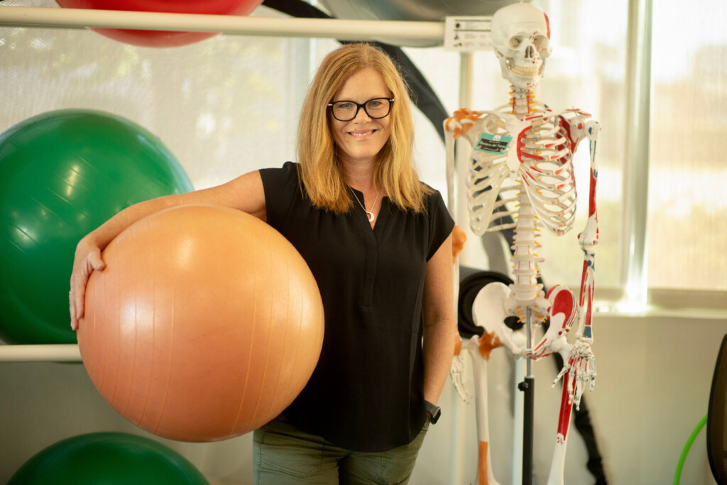 Woman standing in front of work out equipment holding an exercise ball and smiling.