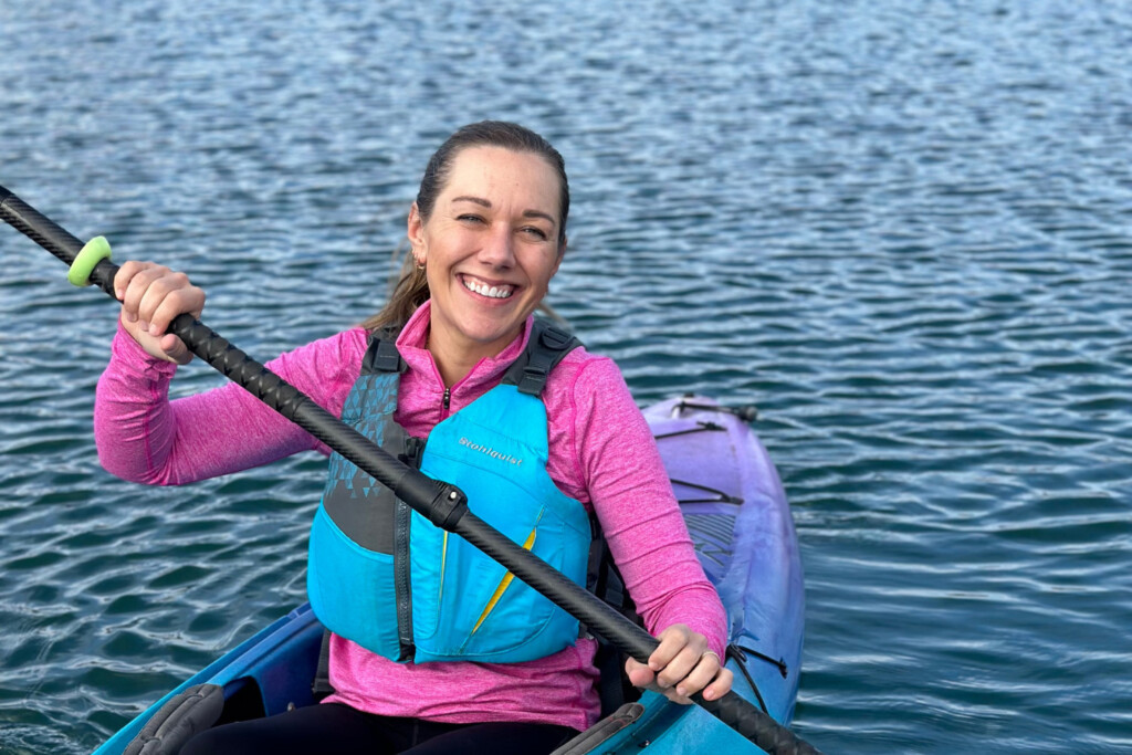 Woman in a kayak on a lake wearing a pink shirt and smiling at the camera.