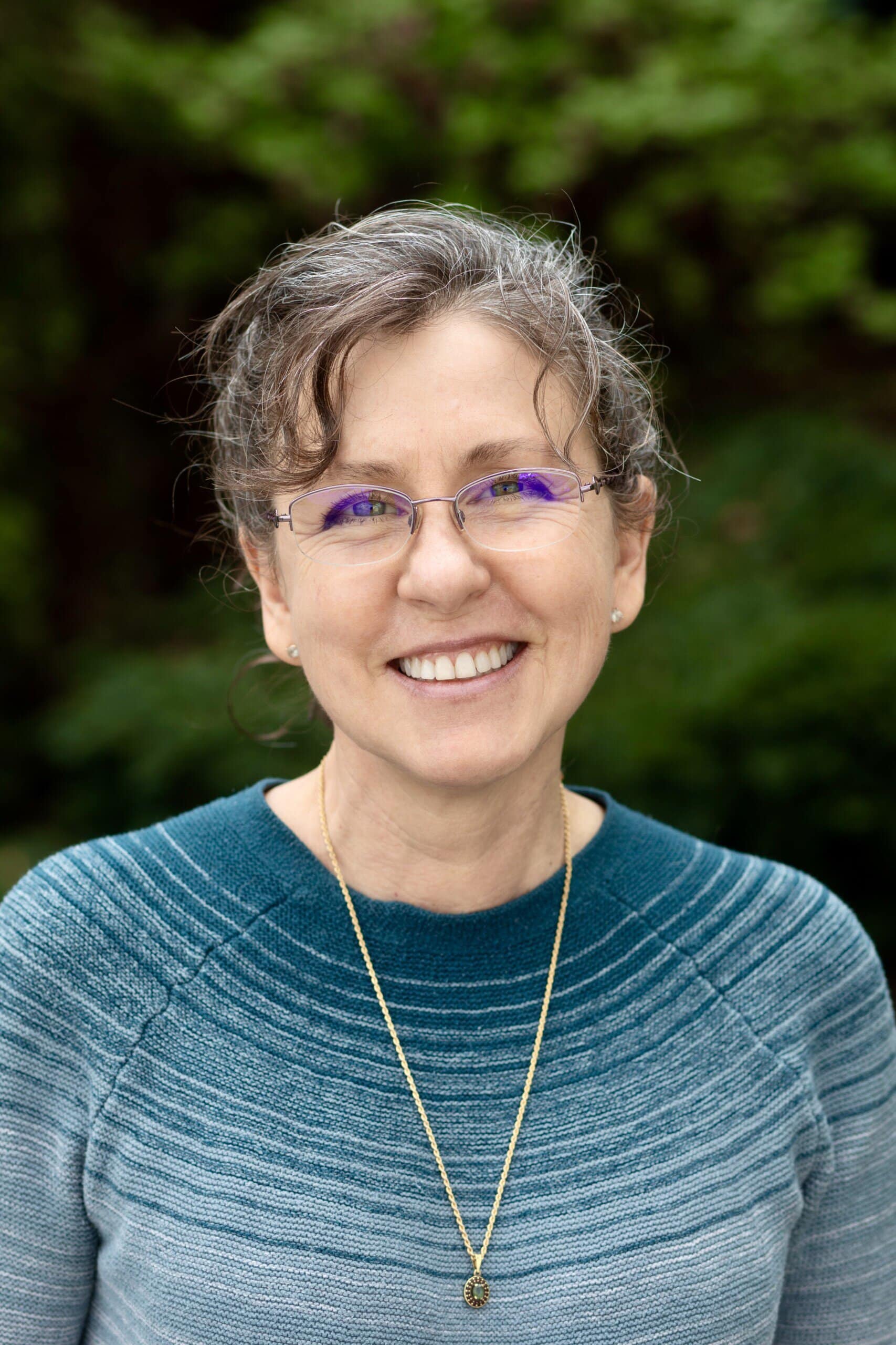 Woman wearing a blue sweater standing in front of green bushes and smiling at the camera.