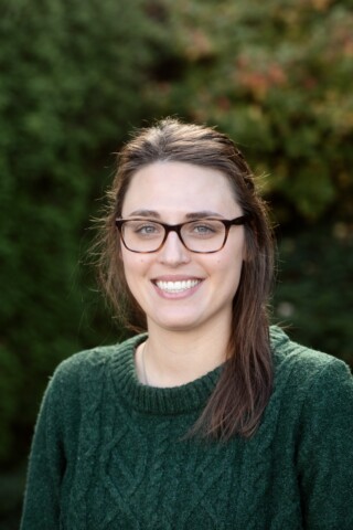 Smiling woman in green sweater, standing in front of green bushes.