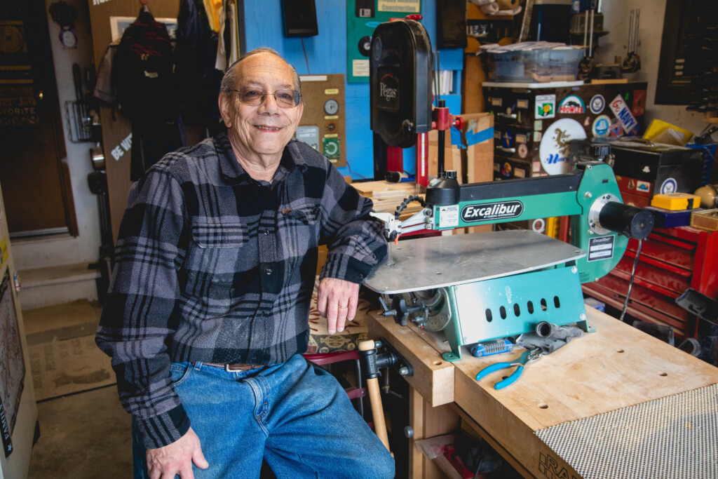 Man in blue and gray flannel shirt sitting at a workbench by a saw in a woodshop.