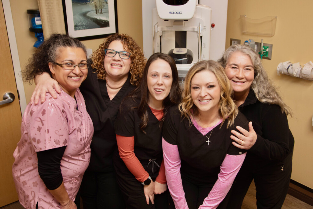 Five women in scrubs standing in front of a mammography machine, smiling.