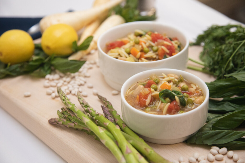 Spring minestrone soup on a cutting board with fresh veggies and lemons around the bowls.