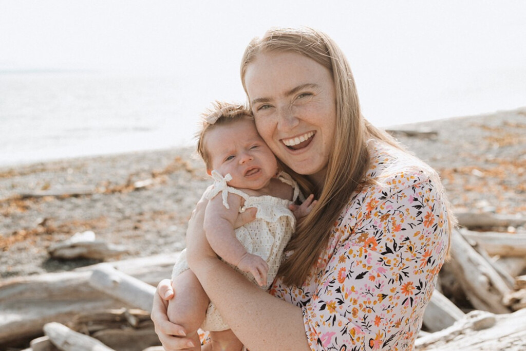 New mother holding newborn smiling while sitting on a beach.