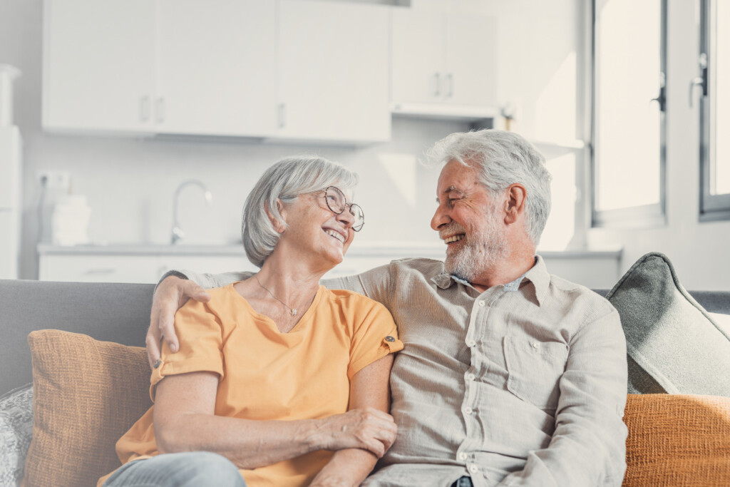 Older couple sitting together on a couch, looking and smiling at each other.