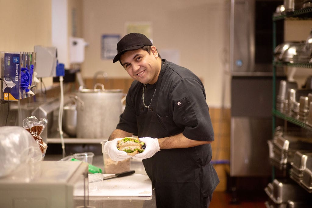 Gus standing in the kitchen holding a tuna fresh-to-go sandwich and smiling.