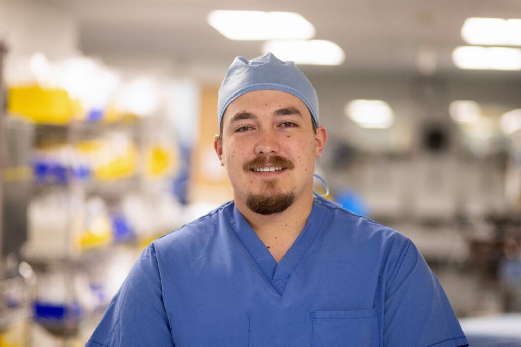 Man in scrubs standing in front of surgical equipment smiling.