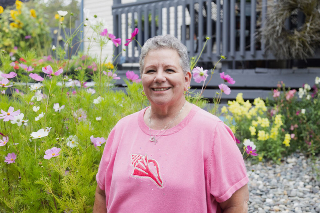 Woman standing in a yard full of flowers wearing a pink breast cancer awareness shirt.