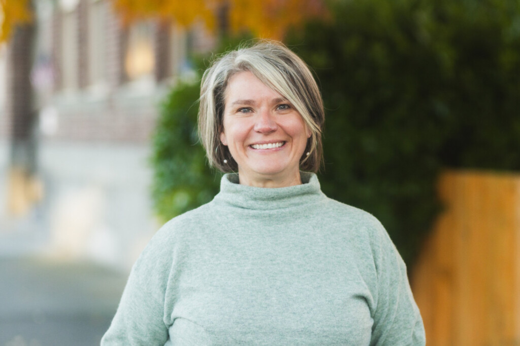 Woman in green sweater standing on a sidewalk smiling.